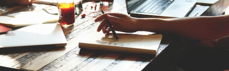 Hands annotating printed spreadsheet reports next to an open laptop on a wooden desk